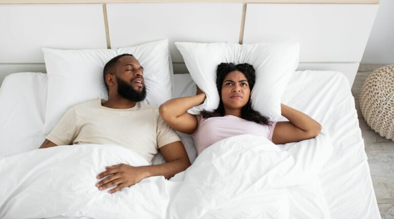 Upset despair millennial black woman covers ears with pillow, suffers from snoring of husband on bed